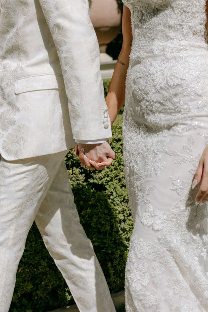 A close-up photo shows a bride in a lace gown holding hands with her groom in a white tux at their wedding at Villa Miani in Rome 