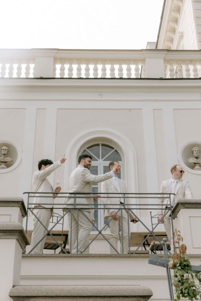 Groomsmen wearing neutrals wave from the balcony at a villa miani rome wedding